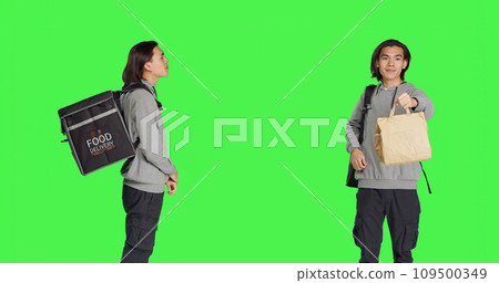 Standing over studio greenscreen backdrop, asian man operates on food delivery with heated bag. Young adult holding backpack for takeout, prepared to give ordered meals to clients. Standing over studio greenscreen backdrop, asian man operates on food delivery with heated bag. Young adult holding backpack for takeout, prepared to give ordered meals to clients. 109500349