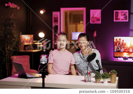Daughter and mother using phone on tripod to film family vlog in home studio, talking about school. Kid and parent recording content for online channel with cellphone camera, discussing with fans Daughter and mother using phone on tripod to film family vlog in home studio, talking about school. Kid and parent recording content for online channel with cellphone camera, discussing with fans 109500358