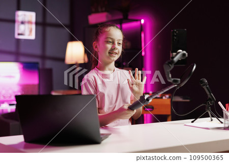 Cheerful young influencer doing livestream using cellphone attached to selfie stick. Lively cute kid does live broadcast using phone camera to talk with online subscribers Cheerful young influencer doing livestream using cellphone attached to selfie stick. Lively cute kid does live broadcast using phone camera to talk with online subscribers 109500365