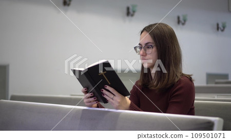 A young woman reads the Bible while sitting on a church bench. A Protestant girl with glasses reads the Bible in church. 109501071