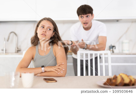 Young girl sitting at the table while her boyfriend is quarreling with her standing behind Young girl sitting at the table while her boyfriend is quarreling with her standing behind 109503646
