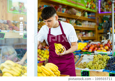 Grocery store employee in an apron puts fresh bananas on shelves Grocery store employee in an apron puts fresh bananas on shelves 109503711