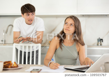 Young girl sitting at the table while her boyfriend is quarreling with her standing behind Young girl sitting at the table while her boyfriend is quarreling with her standing behind 109503767