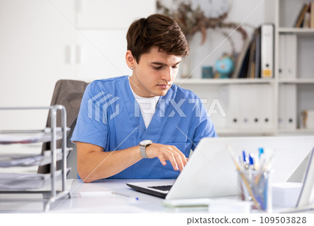 Positive man doctor therapist sitting in office at workplace and working at laptop Positive man doctor therapist sitting in office at workplace and working at laptop 109503828