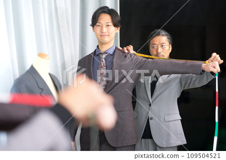Tailor staff taking customer's measurements in front of the mirror Tailor staff taking customer's measurements in front of the mirror 109504521