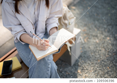 A female is keeping her diary or taking notes in her notebook while resting on a bench in a park. 109506396