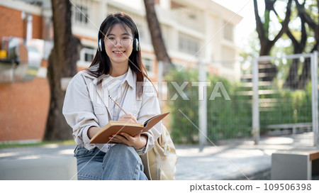 A positive female is listening to music and keeping her diary while resting on a bench in a park. 109506398