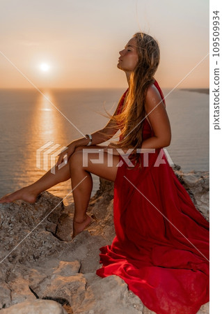 Woman sunset sea red dress, side view a happy beautiful sensual woman in a red long dress posing on a rock high above the sea on sunset. Woman sunset sea red dress, side view a happy beautiful sensual woman in a red long dress posing on a rock high above the sea on sunset. 109509934