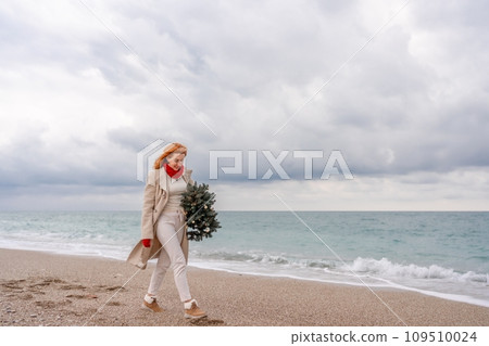 Redhead woman Christmas tree sea. Christmas portrait of a happy redhead woman walking along the beach and holding a Christmas tree in her hands. Dressed in a light coat, white suit and red mittens. 109510024