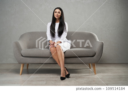 Beautiful long haired female psychotherapist posing to camera, while resting on sofa. Low angle view of elegant woman in medical robe sitting on couch with crossed legs and smiling. Workplace concept. Beautiful long haired female psychotherapist posing to camera, while resting on sofa. Low angle view of elegant woman in medical robe sitting on couch with crossed legs and smiling. Workplace concept. 109514184