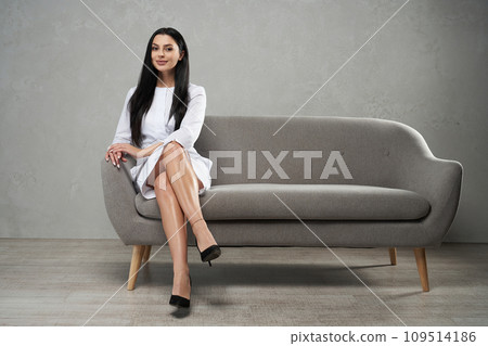 Attractive caucasian female medical staff looking at camera, while sitting on comfortable grey sofa. Front view of smiling woman wearing white robe posing with crossed legs, copy space. Work concept. Attractive caucasian female medical staff looking at camera, while sitting on comfortable grey sofa. Front view of smiling woman wearing white robe posing with crossed legs, copy space. Work concept. 109514186