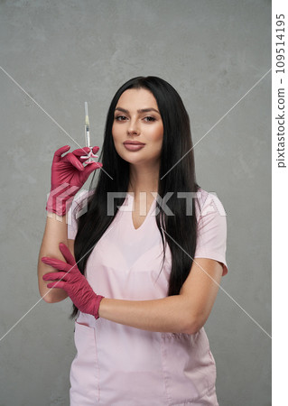 Confident female beautician wearing pink uniform standing with syringe indoors. Portrait of beautiful caucasian woman wearing gloves holding syringe and smiling in clinic. Concept of beauty care. 109514195