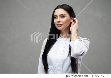 Smiling young woman wearing medical robe, tucking long, dark hair behind ear. Portrait of beautiful caucasian female doctor shyly touching straight hair, while looking away. Concept of emotions, work. 109514205