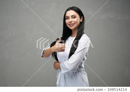 Beautiful female doctor showing thumb up and smiling to camera. Portrait of pretty brunette woman wearing medical uniform giving like gesture, in front of grey background. Medicine, gesture concept. Beautiful female doctor showing thumb up and smiling to camera. Portrait of pretty brunette woman wearing medical uniform giving like gesture, in front of grey background. Medicine, gesture concept. 109514214