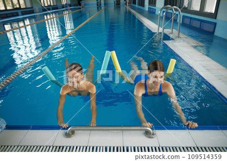 Female coach instructing young woman exercising with pool noodle aerobics class Female coach instructing young woman exercising with pool noodle aerobics class 109514359