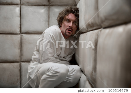 Mad scared man patient wearing straitjacket in corner of padded room 109514479