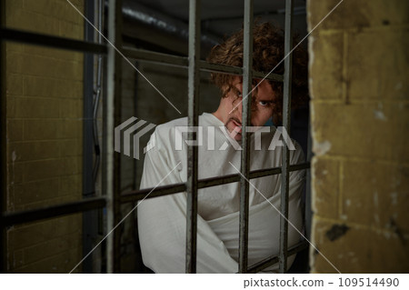 Scared quiet crazy man wearing straightening jacket standing nearby cell gate 109514490