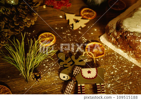 Christmas background with elk toy, dried orange fruit and festive lights. Advent decoration. New Year eve celebration. Christmas card. Winter holidays table. Christmas garland light with sweet stollen 109515188