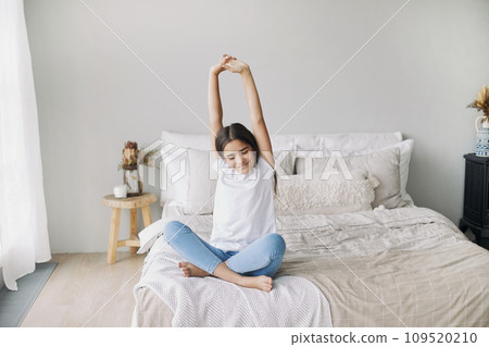 Pre-teen girl in casual clothes do stretching exercises seated on bed in cozy bedroom in the morning 109520210