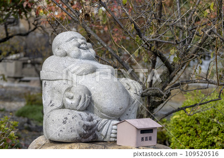 Nagahama, Shiga Prefecture, Japan. Nov 21, 2023. A sitting smiling Buddha stone figure at a Japanese a Shinto shrine. Nagahama, Shiga Prefecture, Japan. Nov 21, 2023. A sitting smiling Buddha stone figure at a Japanese a Shinto shrine. 109520516