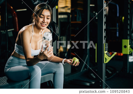 Woman holds a green apple and a water bottle in gym, emphasizing the importance of healthy food choices and staying hydrated for maintaining a healthy lifestyle. Healthy fitness and eating lifestyle 109520843