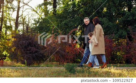 Family with son spends weekend together walking across fall botanical garden. Family of mom and dad leads little son by hands along path with bushes and bench. Family in warm clothes enjoy warm sunset 109522927