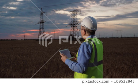 Skilled electrician with helmet analyses data of power distributions substation with tablet in dusk field. Engineer with tablet computer maintains power generation equipment in sunset field 109522945
