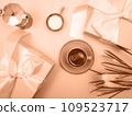 There is a coffee pot on the table, a cup of coffee, a notebook and flowers. The background is peach color. 109523717
