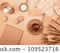 There is a coffee pot on the table, a cup of coffee, a notebook and flowers. The background is peach color. 109523718