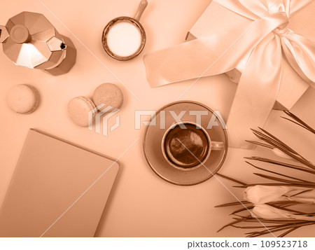 There is a coffee pot on the table, a cup of coffee, a notebook and flowers. The background is peach color. There is a coffee pot on the table, a cup of coffee, a notebook and flowers. The background is peach color. 109523718