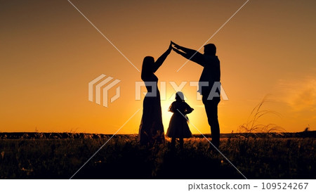 Silhouettes of parents on evening sky holding hands creating home for daughter at sunset 109524267