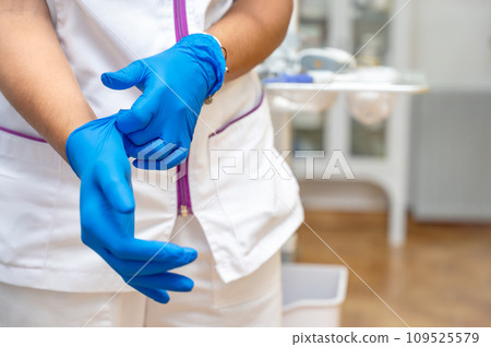 Close up of a female doctor putting a blue latex gloves on a clinic background. Close up of a female doctor putting a blue latex gloves on a clinic background. 109525579