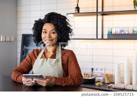 Portrait waitress of a small business owner of a cafe, woman in apron smiles and looks at camera with a tablet computer in her hands, an employee of a cafe of a restaurant near a coffee counter. 109525715