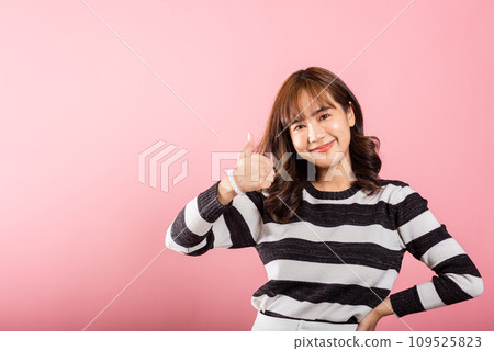 An Asian woman, captured in a studio portrait on pink, expresses agreement and success with a joyful smile and a thumbs-up 'Ok' sign. Her positivity shines through. 109525823