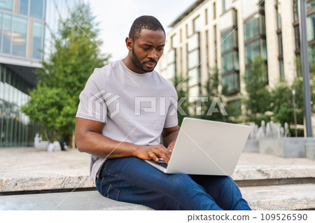 African American man freelancer using laptop typing chatting on urban street in city. Guy having virtual meeting online chat conference. Entrepreneur programmer working on laptop outdoors. Remote work 109526590