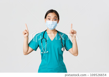 Covid-19, coronavirus disease, healthcare workers concept. Intrigued asian female doctor, physician or intern in medical mask and scrubs, looking curious, pointing fingers up, white background 109526654