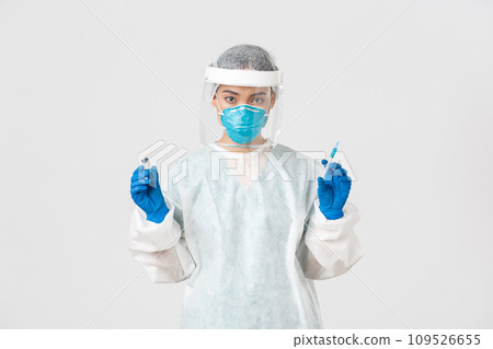Covid-19, coronavirus disease, healthcare workers concept. Confident asian female doctor, physician in personal protective equipment and respirator, ready make ingection, holding syringe with vaccine 109526655