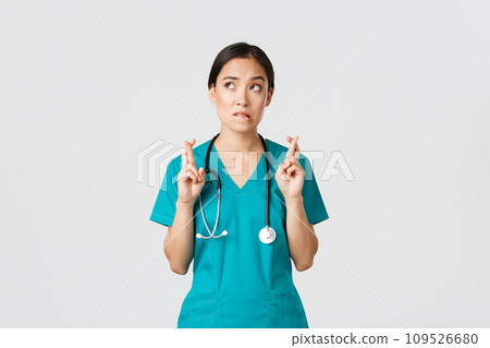 Covid-19, healthcare workers, pandemic concept. Hopeful nervous asian female intern in scrubs waiting for news, doctor cross fingers and biting lip as looking upper left corner, praying 109526680