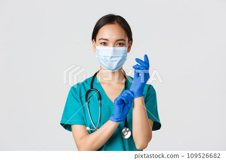 Covid-19, coronavirus disease, healthcare workers concept. Professional smiling asian female physician, doctor in medical mask and scrubs, put on rubber gloves ro examination, white background Covid-19, coronavirus disease, healthcare workers concept. Professional smiling asian female physician, doctor in medical mask and scrubs, put on rubber gloves ro examination, white background 109526682