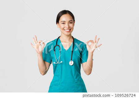 Covid-19, healthcare workers, pandemic concept. Professional confident asian female doctor, intern in scrubs assure patients everything good, showing okay gesture satisfied, smiling pleased 109526724