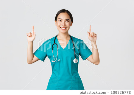 Covid-19, healthcare workers, pandemic concept. Smiling beautiful asian female nurse, doctor in scrubs pointing and looking up with satisfied grin, happy to show awesome promo offer, white background 109526778