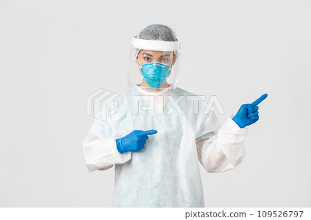 Covid-19, coronavirus disease, healthcare workers concept. Professional female doctor, tech lab employee in personal protective equipment pointing fingers right, showing way, white background 109526797