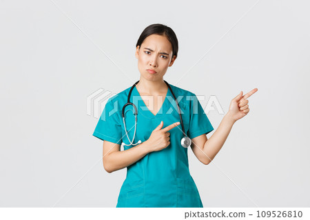 Covid-19, healthcare workers, pandemic concept. Disappointed and angry asian female therapist, nurse in scrubs looking upset at camera, sulking and pointing upper right corner, wait explanation 109526810
