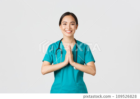 Covid-19, healthcare workers and preventing virus concept. Smiling beautiful asian female nurse, physician in scrubs smiling, hold hands together over chest in namaste, greeting gesture Covid-19, healthcare workers and preventing virus concept. Smiling beautiful asian female nurse, physician in scrubs smiling, hold hands together over chest in namaste, greeting gesture 109526828