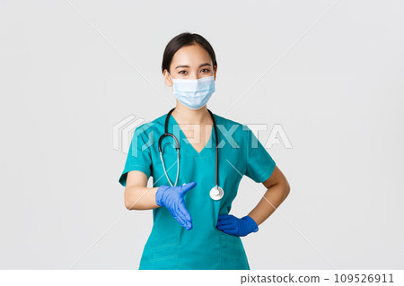 Covid-19, coronavirus disease, healthcare workers concept. Confident young asian female doctor, physician in scrubs and medical mask, gloves, extend hand for handshake, greeting patient 109526911