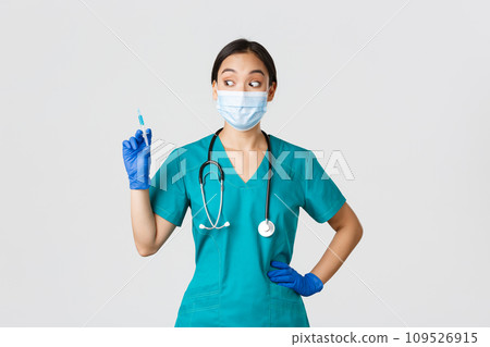 Covid-19, coronavirus disease, healthcare workers concept. Excited asian female nurse in medical mask, rubber gloves and scrubs, looking amazed at syringe with vaccine, white background 109526915