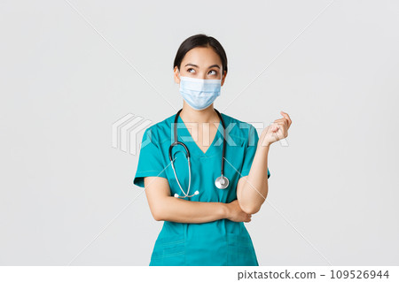 Covid-19, coronavirus disease, healthcare workers concept. Curious and thoughtful asian female doctor in medical mask and scrubs, looking upper left corner, thinking, white background 109526944