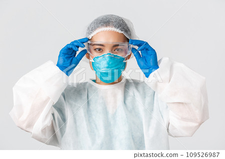 Covid-19, coronavirus disease, healthcare workers concept. Serious-looking asian female doctor in personal protective equipment, put on glass before entering lab, white background 109526987