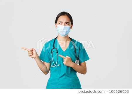 Covid-19, coronavirus disease, healthcare workers concept. Skeptical and disappointed asian female doctor, nurse in scrubs and medical mask looking displeased, cringe aversion, pointing left 109526993
