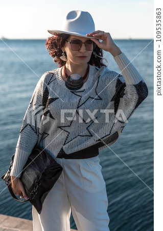 Stylish seashore woman. Fashionable woman in a white hat, white trousers and a light sweater with a black pattern on the background of the sea. 109535863